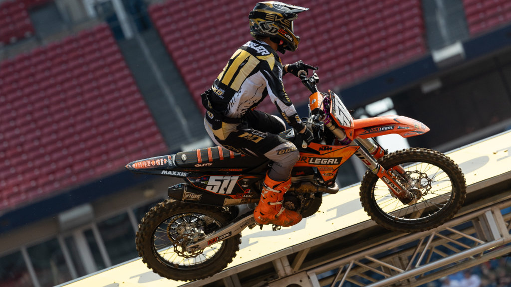 Dirt Legal rider Jace Kessler, number 511, in gold and black gear with orange boots. He is jumping his KTM dirt bike in a stadium, with red arena seats in the blurry background.