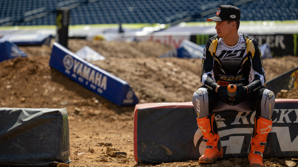 Supercross Rider Relaxing on Track - Dirt Legal A Dirt Legal DBDRacing WMR KTM rider in black, gold, and white gear with orange boots and a hat sits on a track barrier. He looks thoughtfully toward the distance at a Supercross stadium.