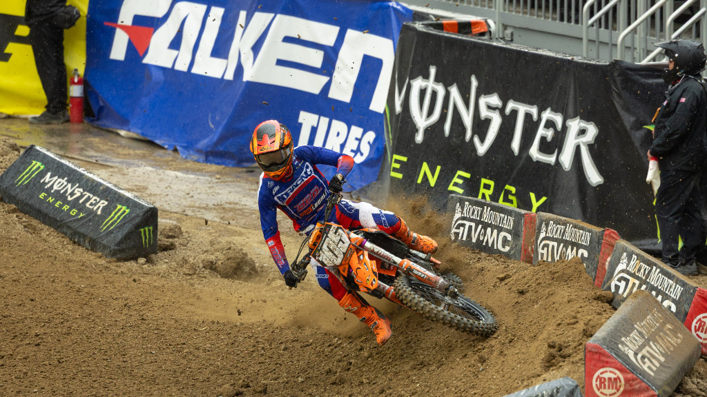 A detailed shot of Marcus Phelps (#105) leaning his mud-caked Dirt Legal DBDRacing KTM dirt bike hard into a left-hand turn on a rainy track.