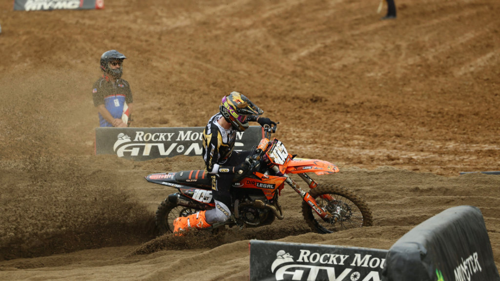 Dirt Legal rider Marcus Phelps in action, wearing gold and black gear with bright orange boots. He is power sliding his orange KTM dirt bike through deep sand on a race track, kicking up a large roost of dirt and sand.