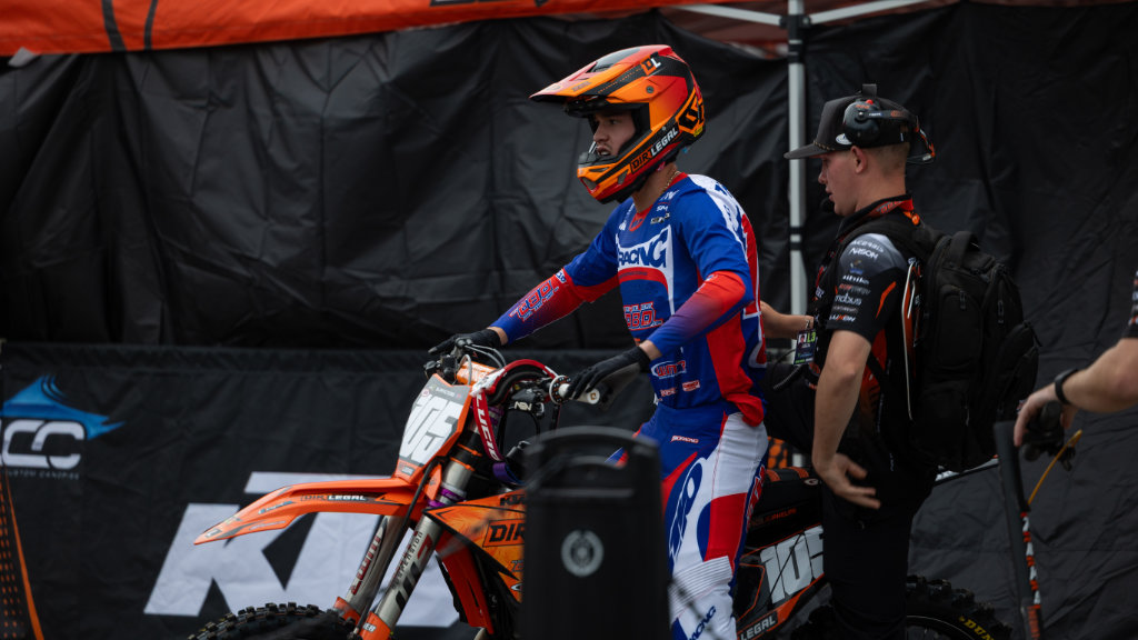 Marcus Phelps (rider #105) seated on his KTM 250 in the team pits. He is wearing his Dirt Legal DBDRacing jersey and helmet, looking forward with concentration.