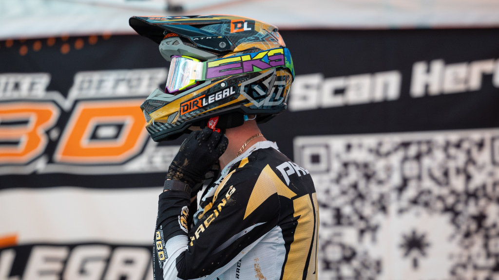 A close-up of a motocross rider in black and gold gear, adjusting a custom, colorful 6D helmet featuring "Dirt Legal" and "EKS" logos before a race.