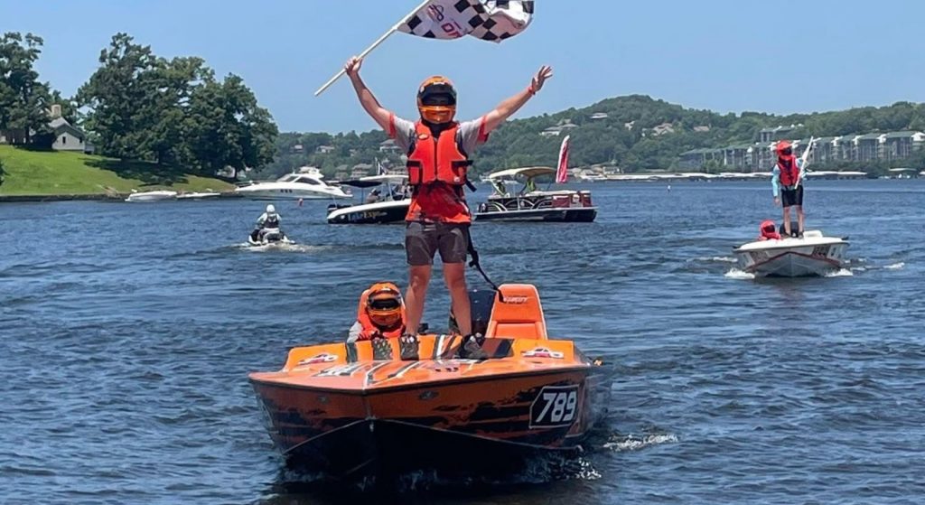 Alt Text: Young racer celebrating on an orange Dirt Legal offshore race boat during a boating event on the lake.
