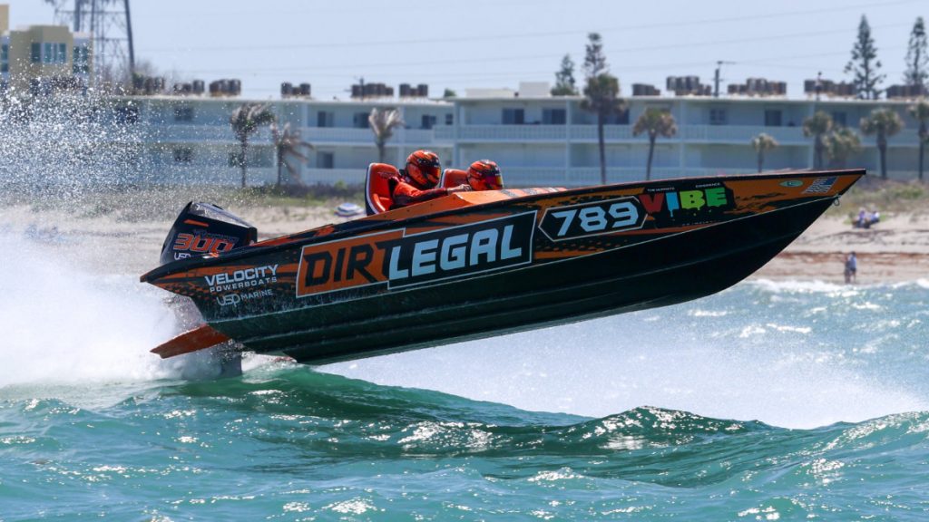 Dirt Legal Velocity Powerboats offshore race boat #789 launching off a wave at Cocoa Beach, with both crew members in orange helmets and the Dirt Legal livery fully visible