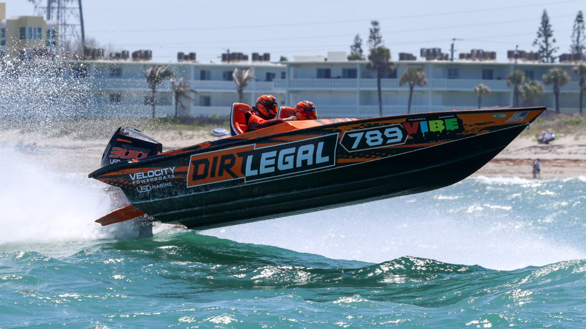 Dirt Legal Velocity Powerboats offshore race boat #789 launching off a wave at Cocoa Beach, with both crew members in orange helmets and the Dirt Legal livery fully visible