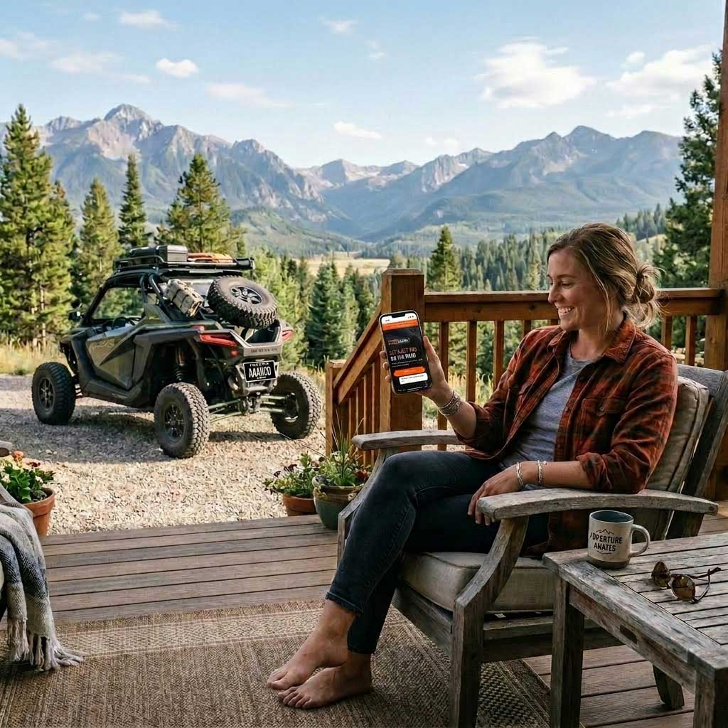 Woman sits barefoot on a cabin deck, smiling at her phone with an ATV parked outside and mountains in the background.