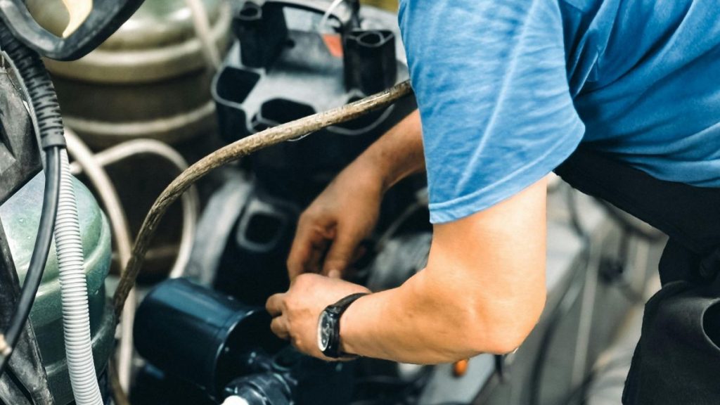 A mechanic in a blue shirt works intently on a complex engine system, adjusting components and hoses inside a workshop setting.
