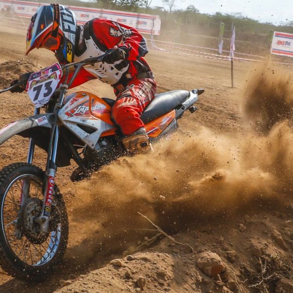 A motocross rider in full protective gear leans aggressively into a dirt track turn, kicking up a massive cloud of dust during an intense off-road race.