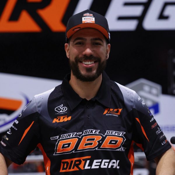 A portrait of professional racer Anthony Rodriguez wearing a Dirt Legal DBDRacing team shirt and hat, smiling while standing behind his KTM race bike.