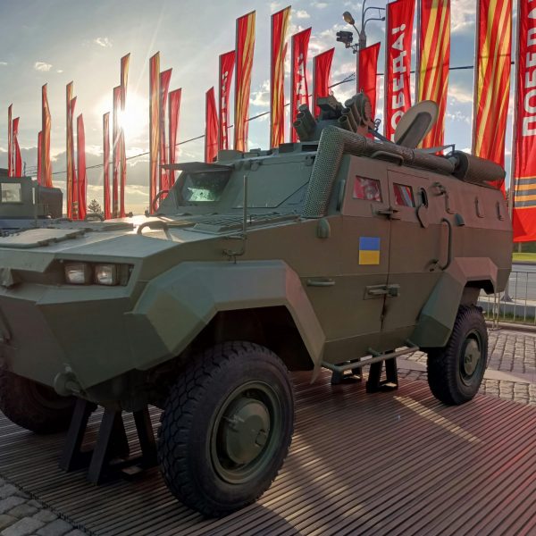 An armored military vehicle is displayed outdoors on a paved platform, finished in matte green with rugged tires, roof-mounted equipment, and national insignia, set against a backdrop of red event banners.