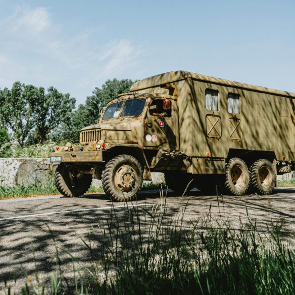 A vintage military cargo truck with a canvas-covered rear drives along a quiet road, surrounded by trees and greenery, showcasing its rugged, utilitarian design.